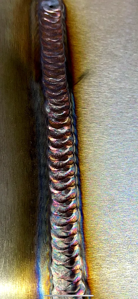 Copper-toned TIG weld bead with even ripples running along brushed stainless steel