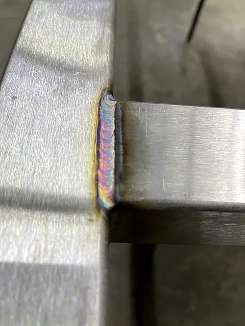 Close-up of a TIG weld with rainbow heat tint joining stainless steel square tubing to a flat bar