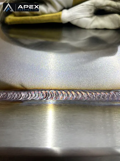 Horizontal stacked-dime TIG weld across stainless steel plates, welding gloves resting nearby