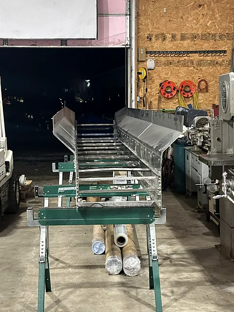Long stainless steel trough conveyor section supported on stands inside the fabrication shop
