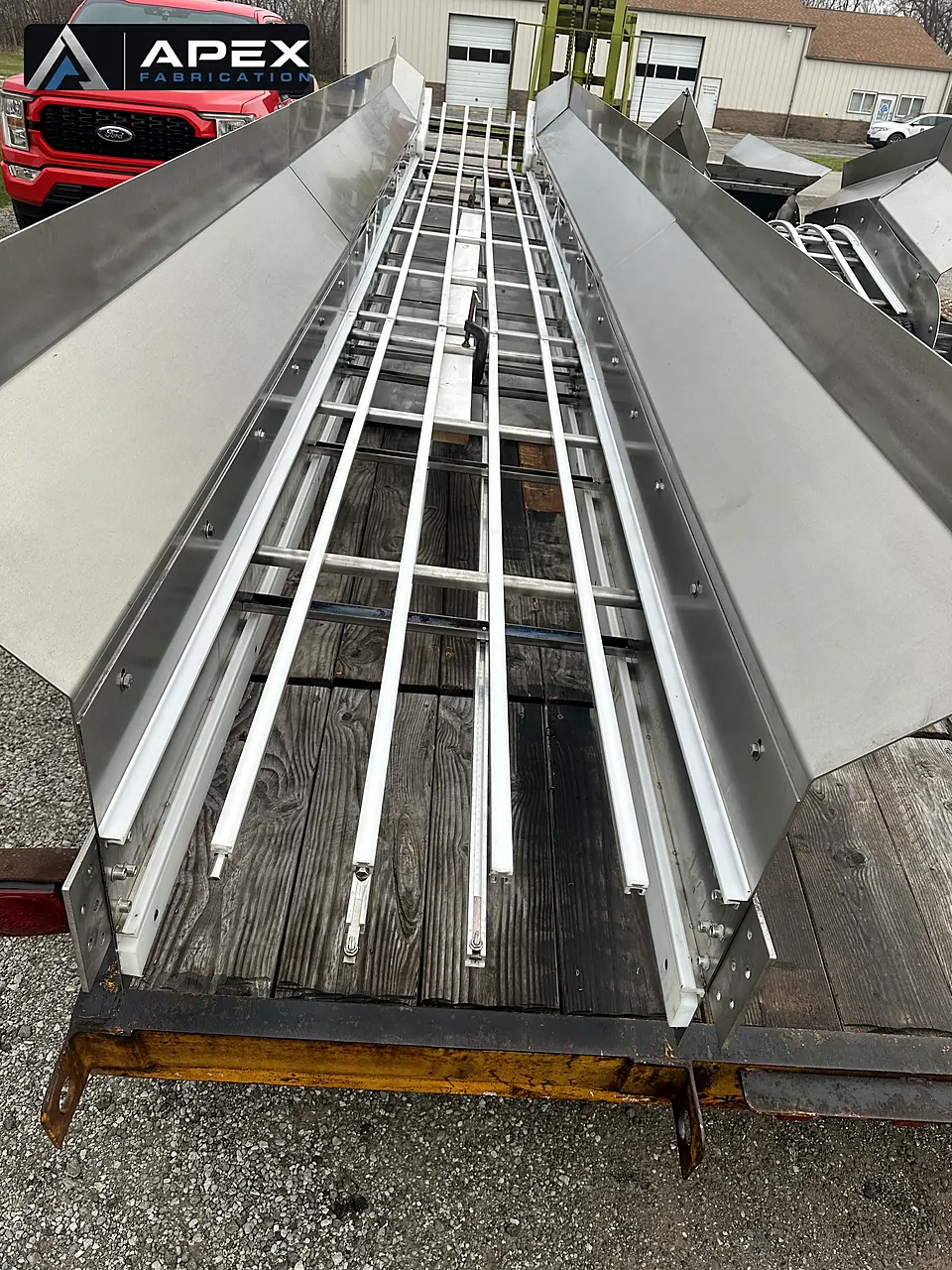 Two stainless steel conveyor trough sections with guide rails laid out on a wooden trailer deck